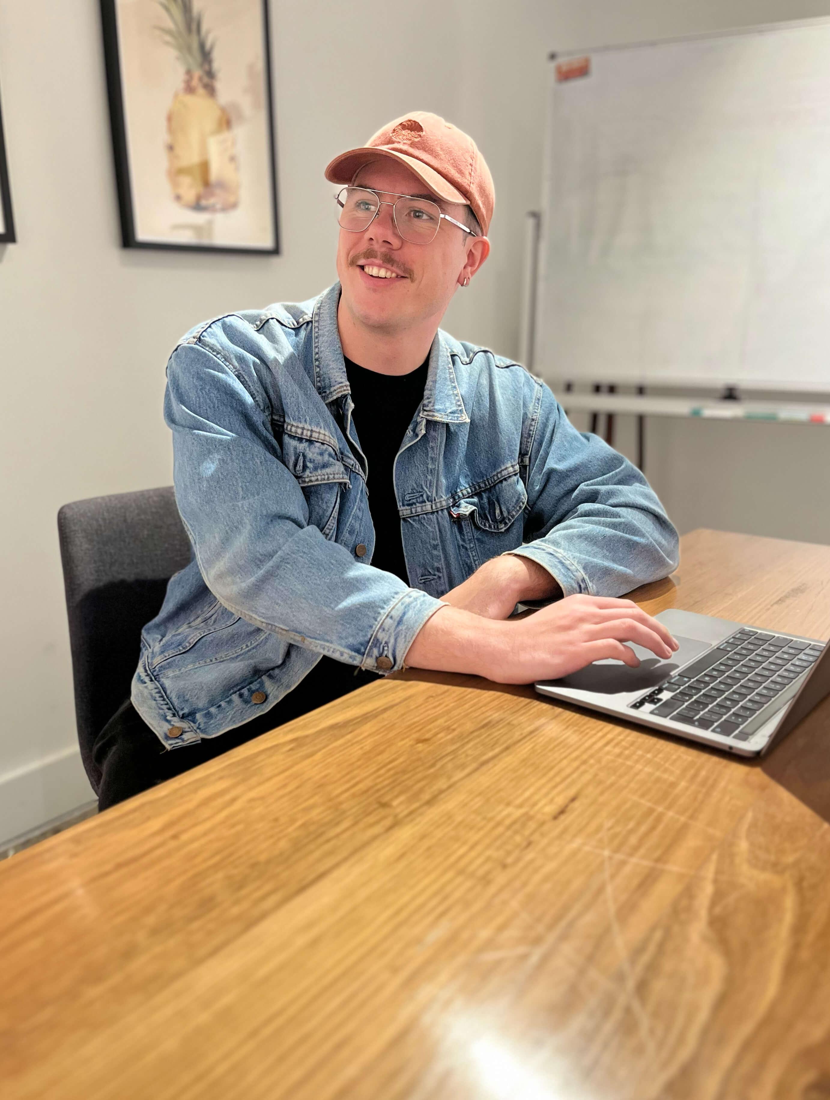 Jack Christian sat at a wooden desk with his laptop open smiling to someone off camera